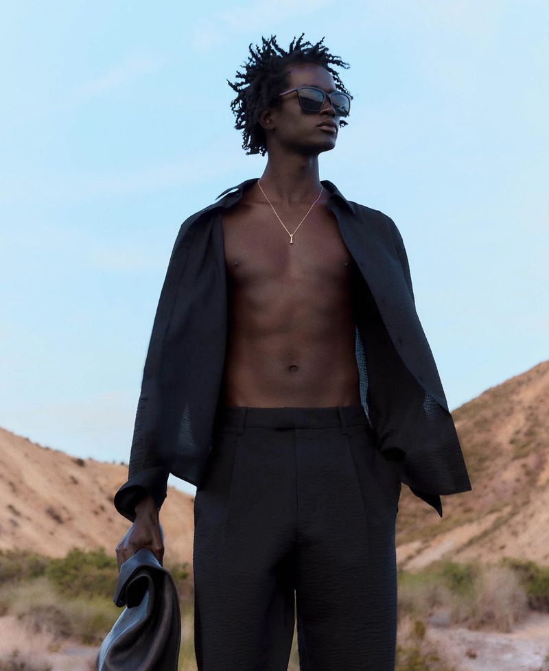 A black male model stands outdoors in a black open dress shirt, black slacks, and sunglasses against a desert backdrop on a sunny day.