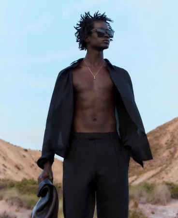 A black male model stands outdoors in a black open dress shirt, black slacks, and sunglasses against a desert backdrop on a sunny day.