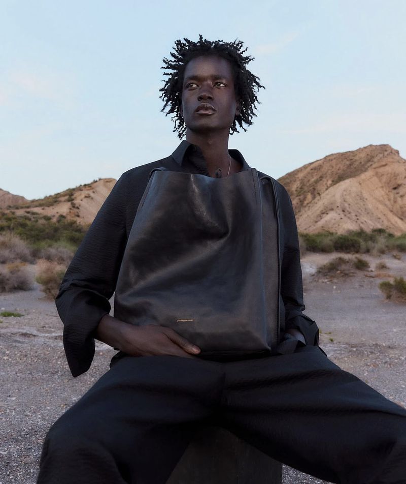 Dark-skinned male model wears a black shirt and pants, sitting with a black leather bag, set against a desert background.