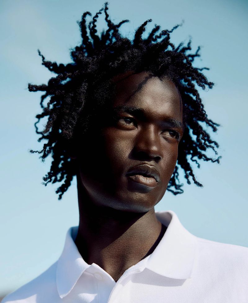 Portrait of a Black man in a white polo shirt against a bright blue sky, showcasing his dark skin, tightly curled hair, and neutral expression.