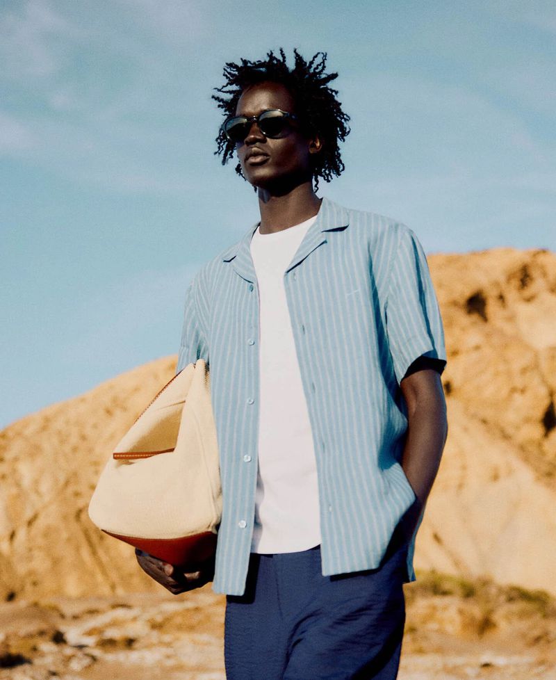 A slender adult male with a tote bag standing outside. He wears sunglasses, a short-sleeved shirt, t-shirt, and pants in a sunny outdoor setting.