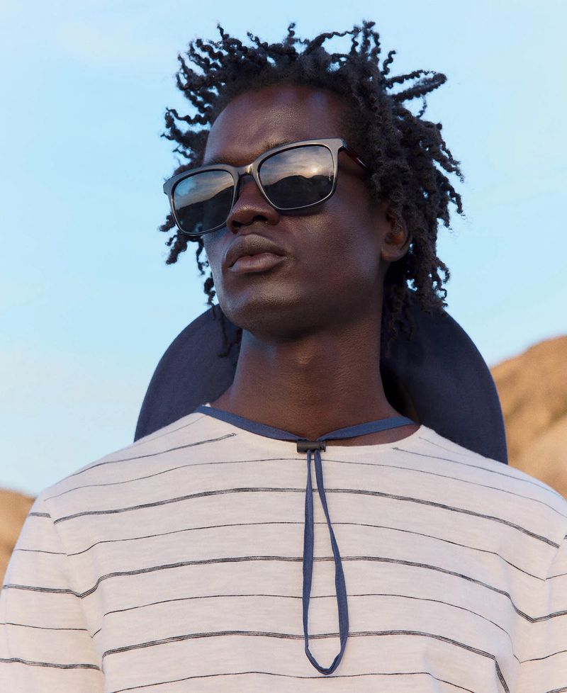 Portrait of an adult male with dark skin and dreadlocks, wearing sunglasses and a striped shirt, posing outdoors against a sky background.