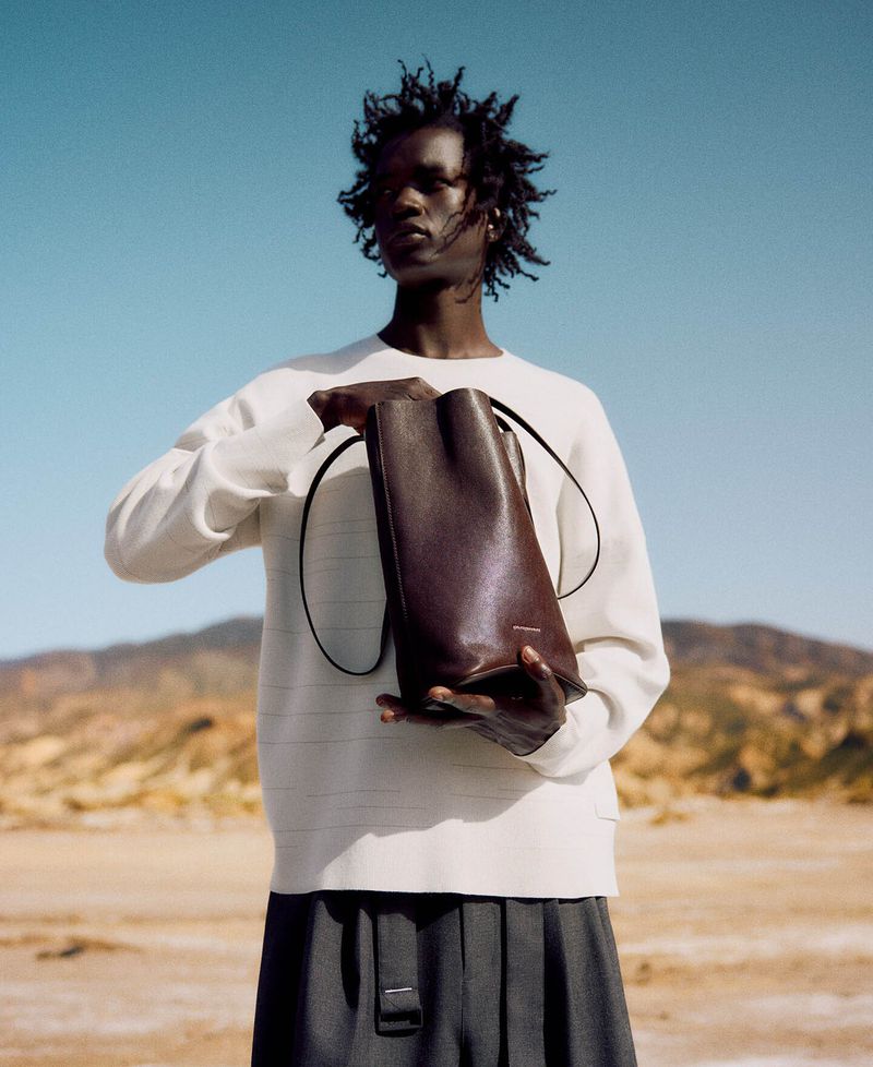 Fashion shot of a Black male model holding a leather tote bag in a desert landscape wearing a white long-sleeve shirt and grey pleated pants.