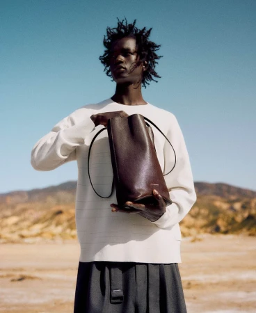 Fashion shot of a Black male model holding a leather tote bag in a desert landscape wearing a white long-sleeve shirt and grey pleated pants.