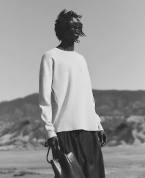 Black man wearing sunglasses and a white sweater holding a leather tote bag outdoors against a mountain background.