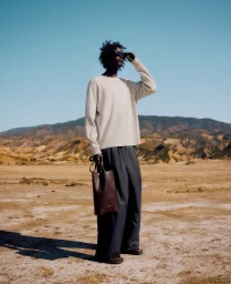 Model wearing sunglasses, a light knit sweater, and dark trousers holding a leather bag in a desert landscape