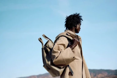 Black man wearing a beige jacket and carrying a backpack against a clear blue sky background