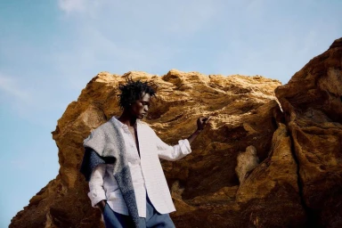 A man in a white button-down shirt and blue trousers stands against a rocky cliff under a clear sky.