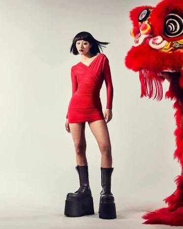 Woman in red dress poses next to traditional Chinese lion dance headpiece against a white background, wearing platform boots.