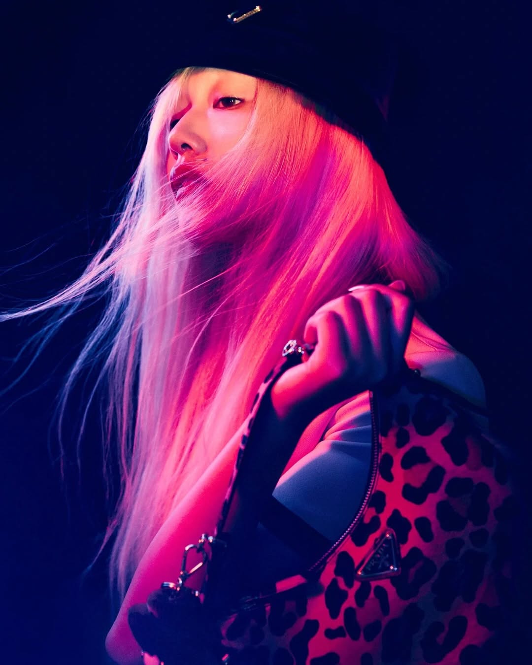 Blonde woman in beret poses with leopard-print Prada purse in a studio setting with dark background and pink and blue lighting.