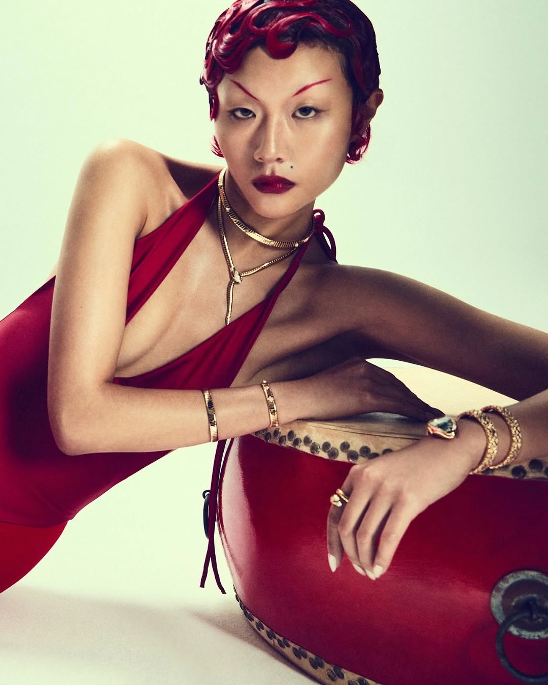 Woman in red dress poses with drum and jewelry. Model with crimson hair and lips in a fashion or beauty context indoors.
