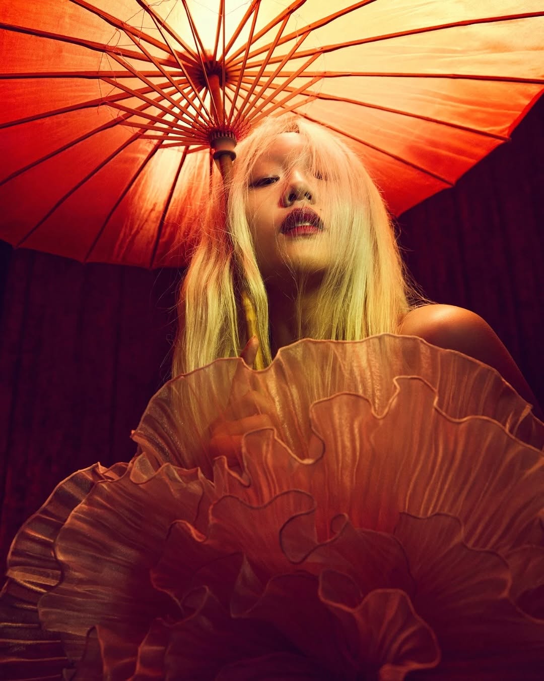 Artistic portrait of a woman with blonde hair under an umbrella. She is wearing a ruffled top and has a neutral facial expression.