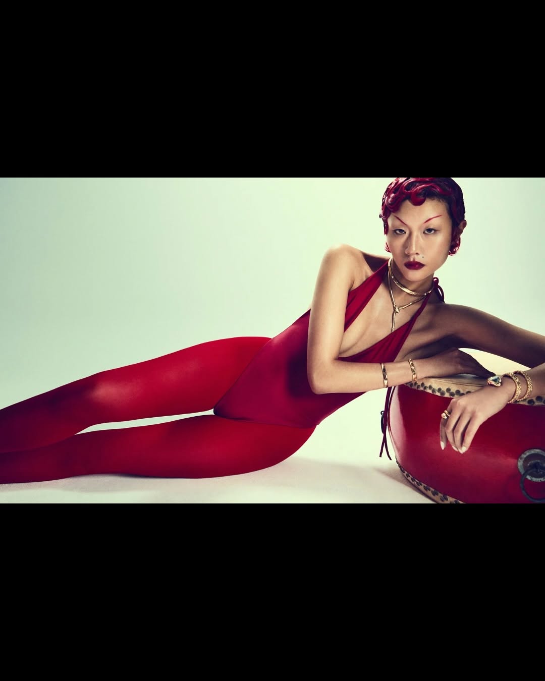 Asian woman in red bodysuit poses with a red drum in a fashion-style studio shot with vibrant colors and a contemporary aesthetic.