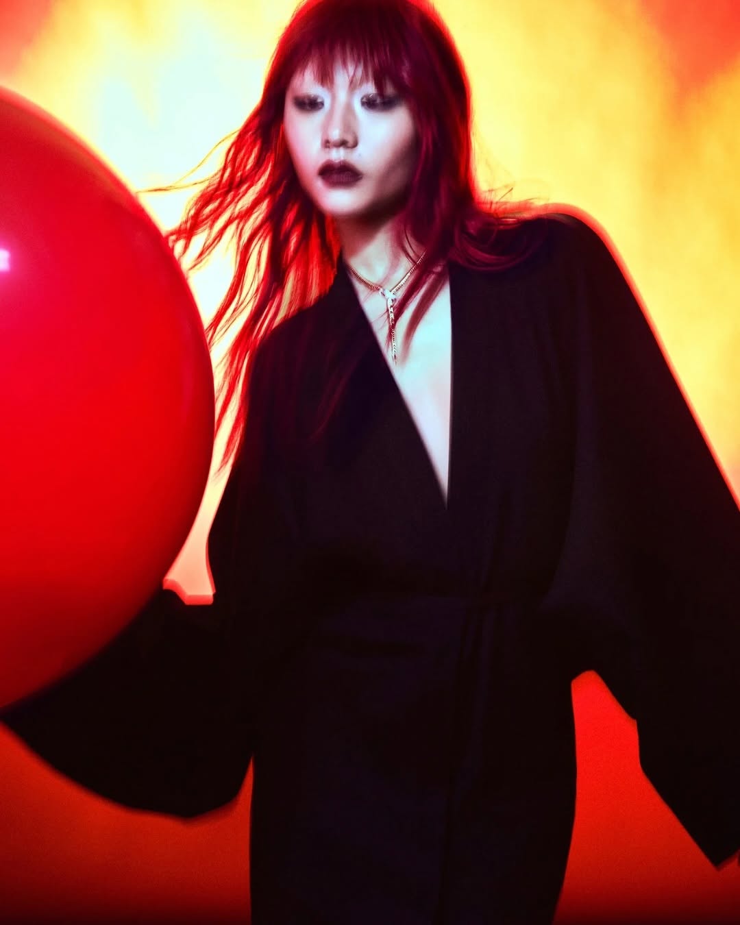 Portrait of a female model with red hair wearing a black blazer next to a large red balloon in a studio setting with warm lighting.