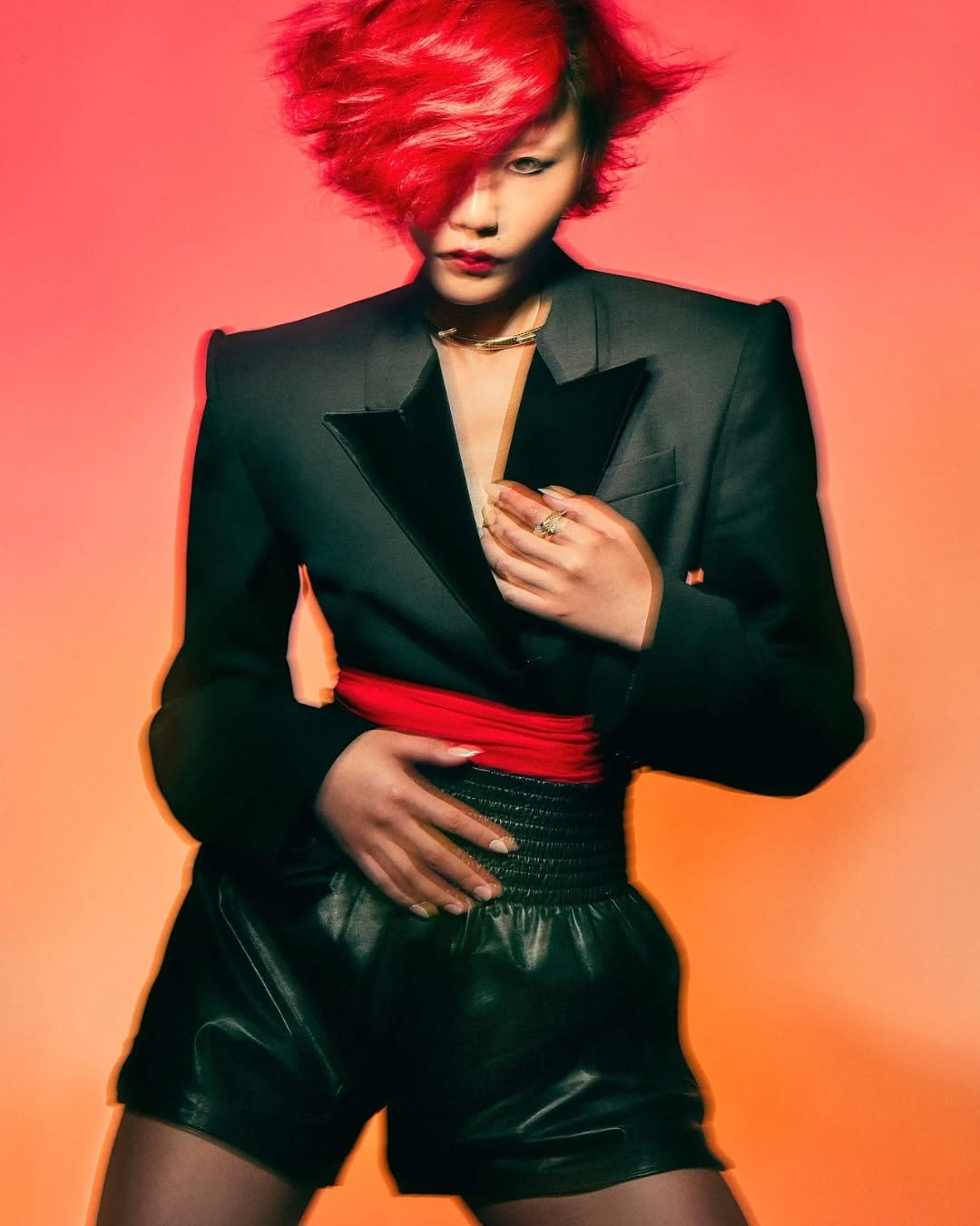 Portrait of a stylish young adult with red hair, wearing a black blazer and leather shorts against a gradient orange background.