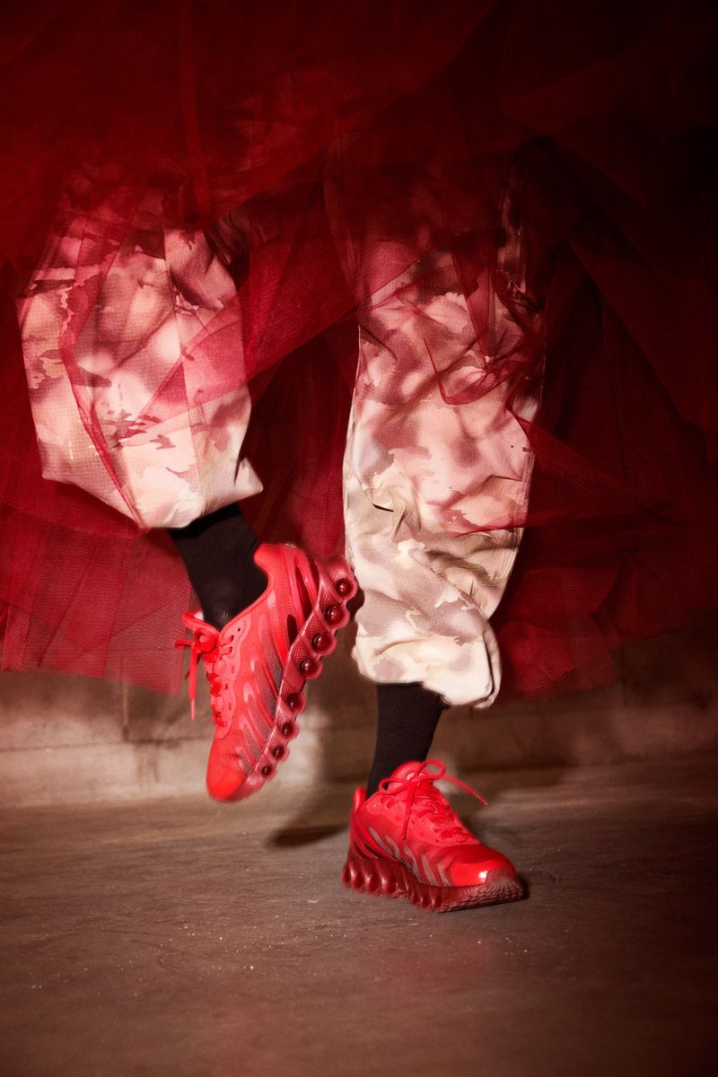 Eye-level studio shot of a person wearing red athletic shoes and patterned pants, with a red background and neutral concrete floor.