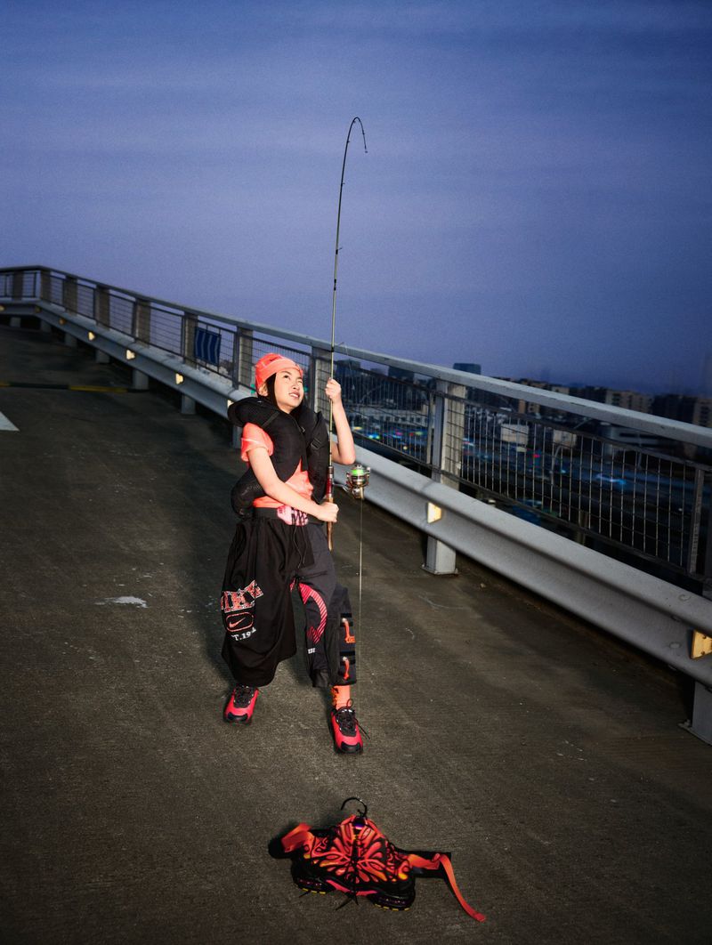 Woman in streetwear fashion poses on a rooftop at night, holding a fishing rod. A red floral bag rests on the concrete.