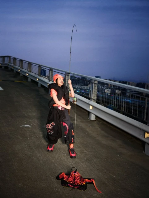 Woman in streetwear fashion poses on a rooftop at night, holding a fishing rod. A red floral bag rests on the concrete.