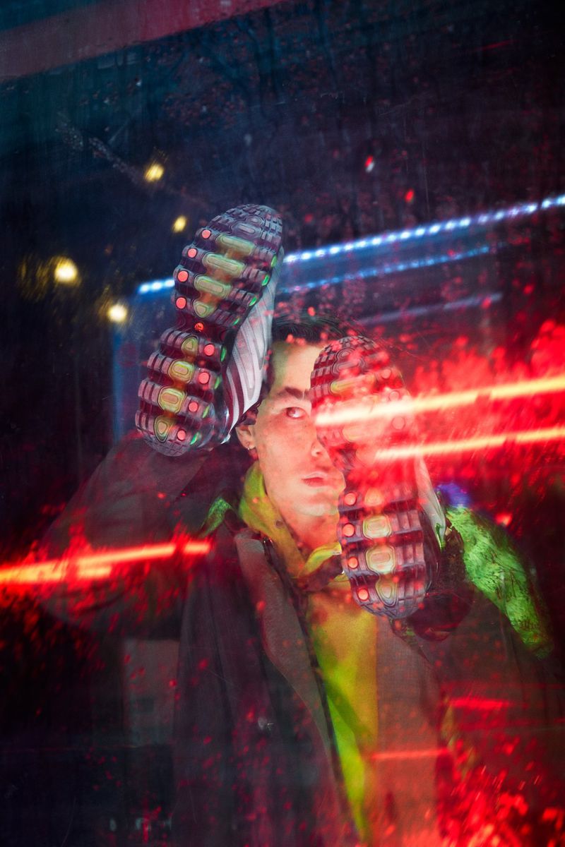 Urban fashion: A man in a coat and bright hoodie poses indoors with a sneaker sole visible, reflected in a wet window with neon lights.
