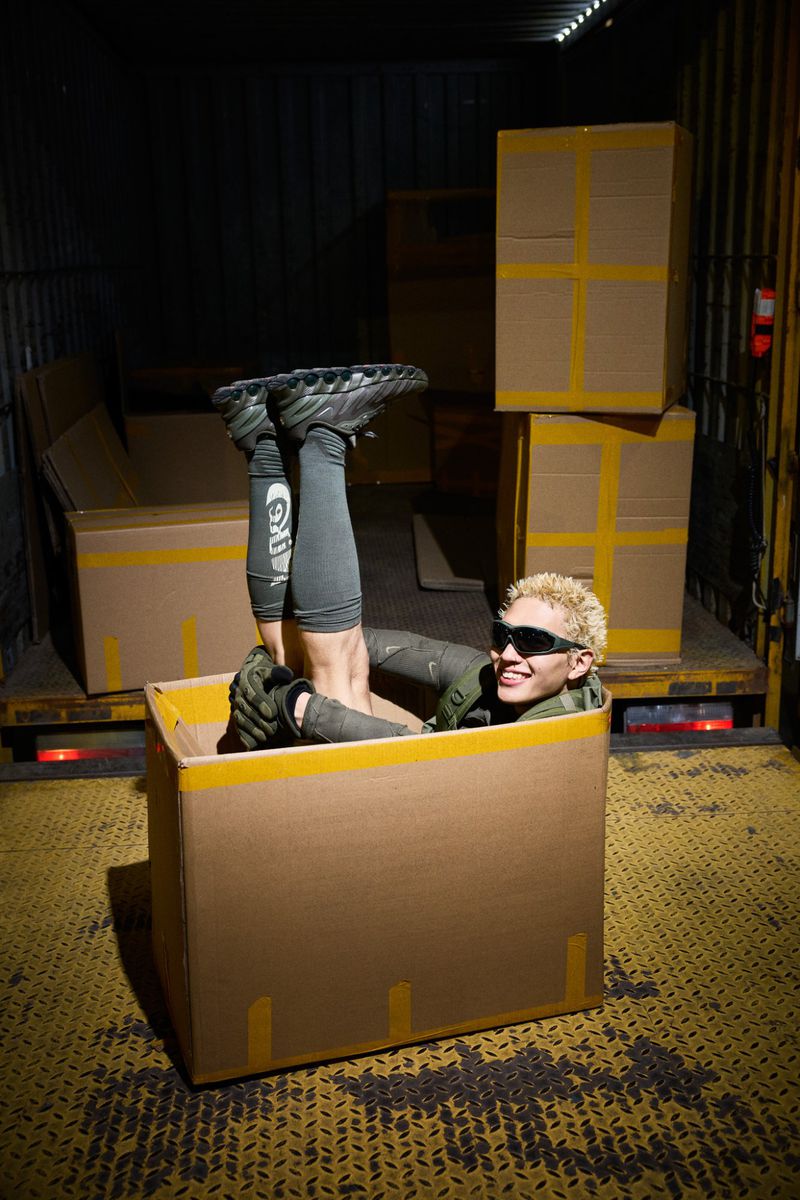 Person with bleached hair in a cardboard box inside a truck, wearing sunglasses, with legs up and feet in the air. 
