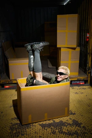 Person with bleached hair in a cardboard box inside a truck, wearing sunglasses, with legs up and feet in the air. 