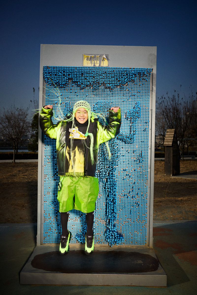 Young person in vibrant neon green and black outfit jumps in front of interactive pin impression wall outdoors on a sunny day.