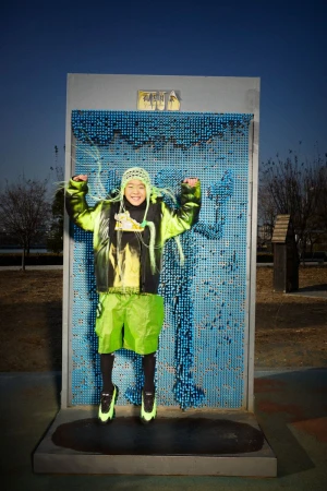 Young person in vibrant neon green and black outfit jumps in front of interactive pin impression wall outdoors on a sunny day.