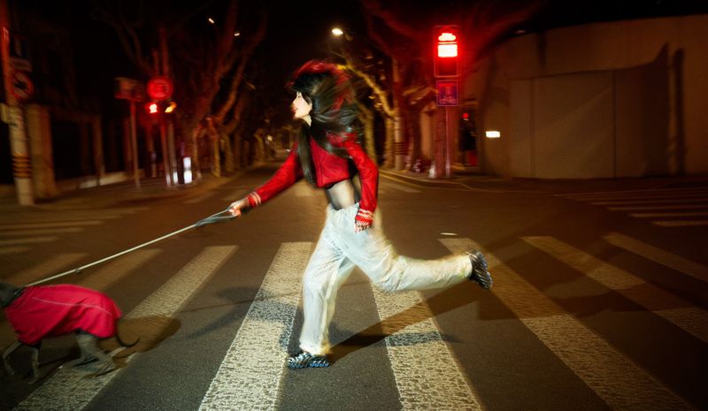 An adult woman runs across a crosswalk at night with a dog in a red coat, streetlights and city buildings lining the background.