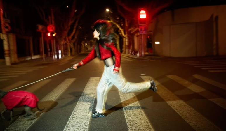 An adult woman runs across a crosswalk at night with a dog in a red coat, streetlights and city buildings lining the background.