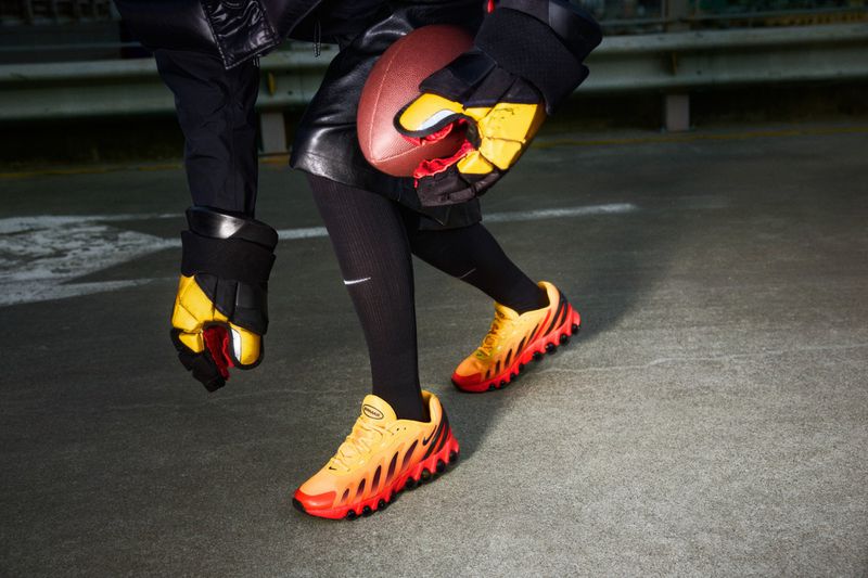 Person wearing black and yellow football gloves and orange sneakers holding an American football on a gray surface.