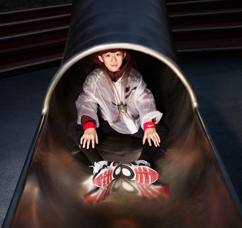 Young child in clear jacket enjoys sliding down a metal tube slide at a playground, captured in a close-up with soft lighting.