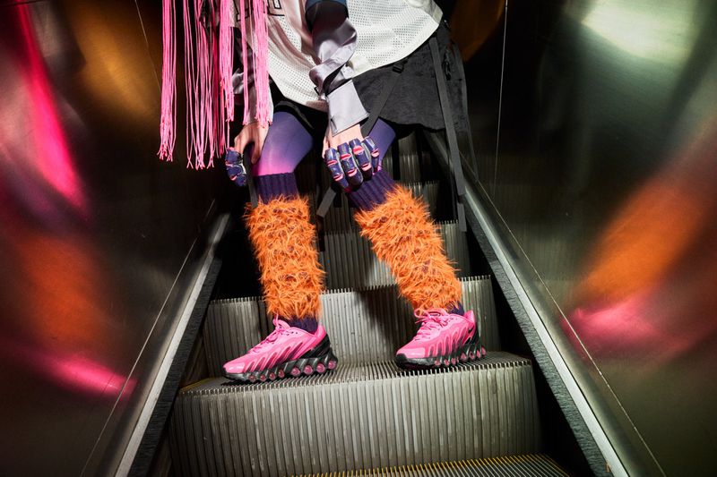 Person stands on an escalator wearing pink sneakers, fuzzy orange leg warmers, and a white jersey with pink hair extensions.