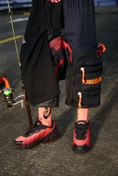 Close-up of Nike Air Max DN8 sneakers in red and black paired with printed socks and utility shorts on a paved surface.