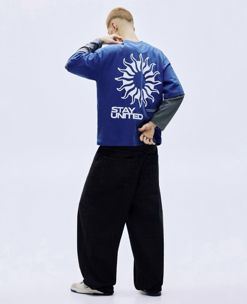 Back view of a person wearing a blue long-sleeve shirt with a white sun graphic and the words 'STAY UNITED,' paired with black pants.