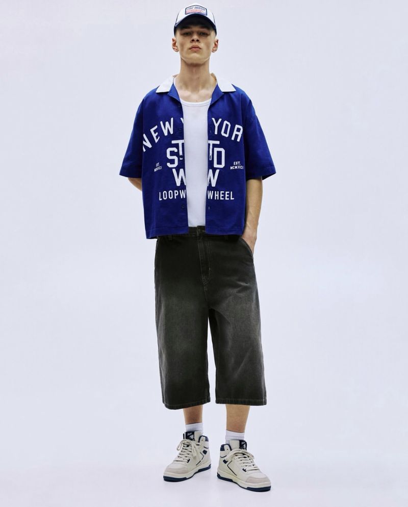 A young adult male poses in a studio wearing a blue camp shirt, white t-shirt, capri pants, and high-top sneakers against a white background.