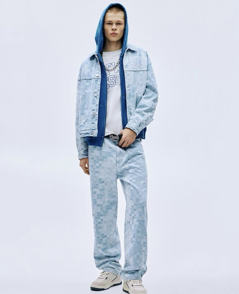 Adult male fashion model posing in a light blue checkerboard print denim jacket and pants outfit in front of a white background.