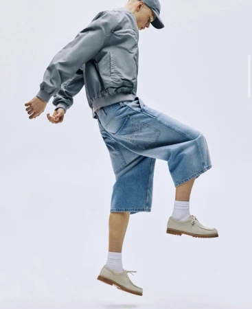 Man wearing blue bomber jacket, baggy denim shorts, white socks, and casual shoes poses mid-air against a bright white backdrop.