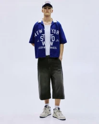 Model wearing a blue graphic bowling shirt, dark jorts, and a baseball cap against a plain white studio background.