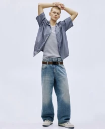Young male model posing in striped short sleeve shirt, grey t shirt, and baggy denim jeans against a white background