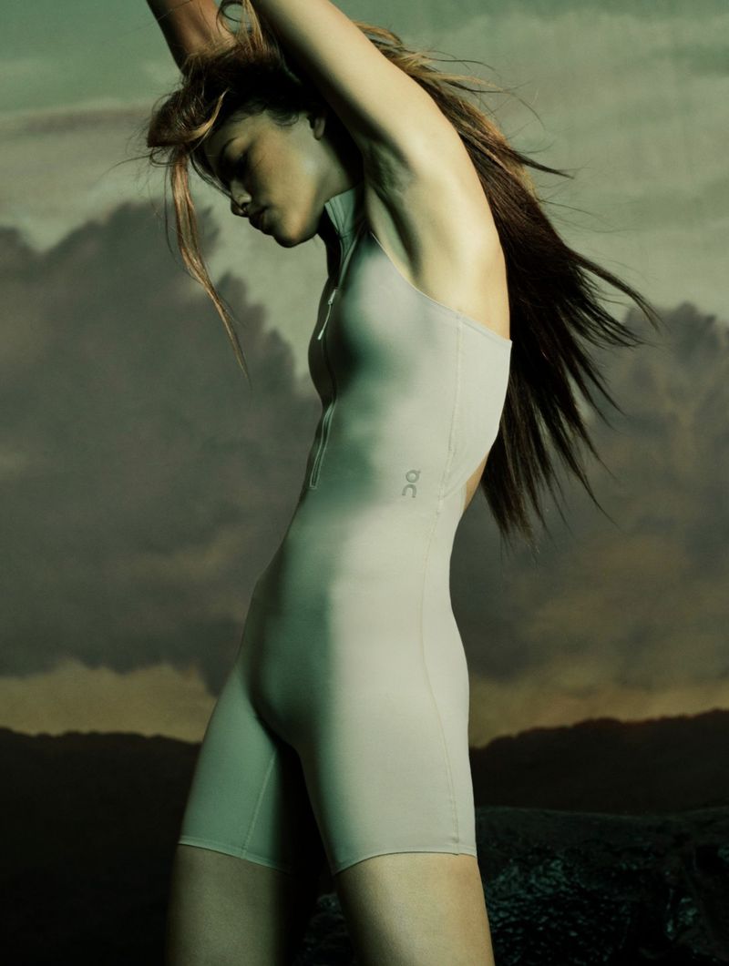 Woman with long brown hair poses in a form-fitting gray bodysuit against a background of dark storm clouds and low mountain range.