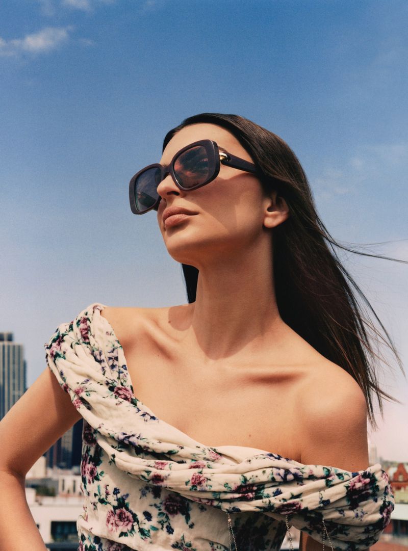 A woman in sunglasses and a floral dress stands against a blue sky and urban backdrop, presenting fashion and style outdoors.