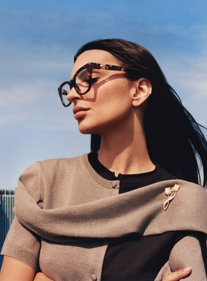Adult woman in a half-tone cardigan and tortoiseshell glasses poses against a blue sky.  She has dark hair and light skin.