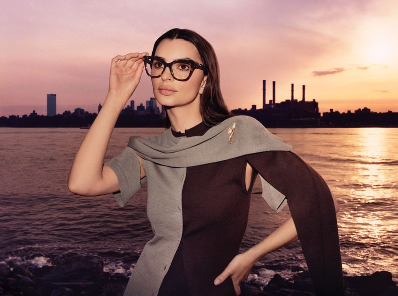 A woman with glasses poses outdoors near a river at dusk, wearing a two-tone grey and brown dress with a cityscape and sunset in the background.