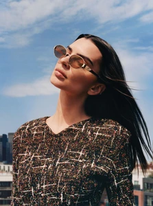Emily Ratajkowski wearing oval sunglasses and a patterned tweed top outdoors against a blue sky with distant buildings.
