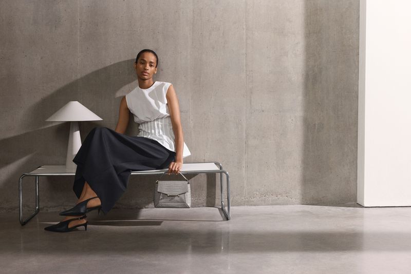 A woman in a white top and black skirt sitting on a bench with a handbag, next to a modern lamp against a gray studio wall.