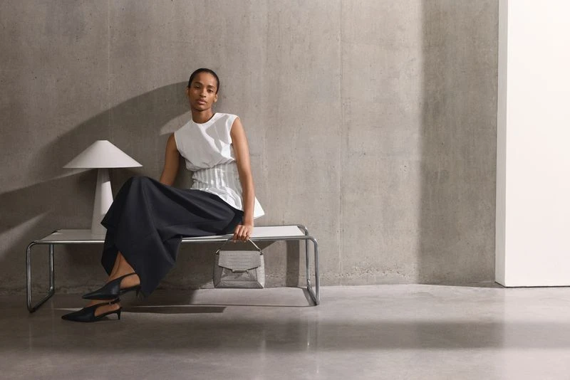 A woman in a white top and black skirt sitting on a bench with a handbag, next to a modern lamp against a gray studio wall.