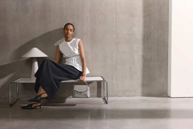 A woman in a white top and black skirt sitting on a bench with a handbag, next to a modern lamp against a gray studio wall.