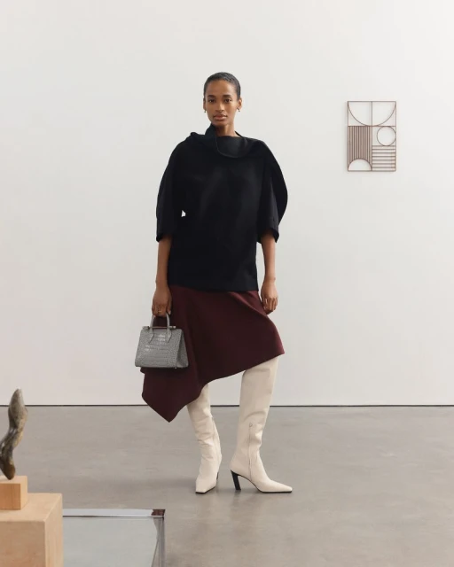 Fashionable adult woman wearing a black top, burgundy skirt, and high boots stands in a studio holding a grey handbag.