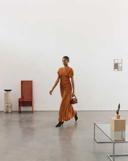 Woman in orange dress walks across gallery, carrying a small handbag; neutral backdrop with modern furniture and artwork on the wall.