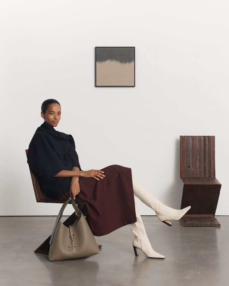 Adult female seated in a modern chair in a light grey room, wearing a black top, dark-red skirt, and white boots; holding a bag.
